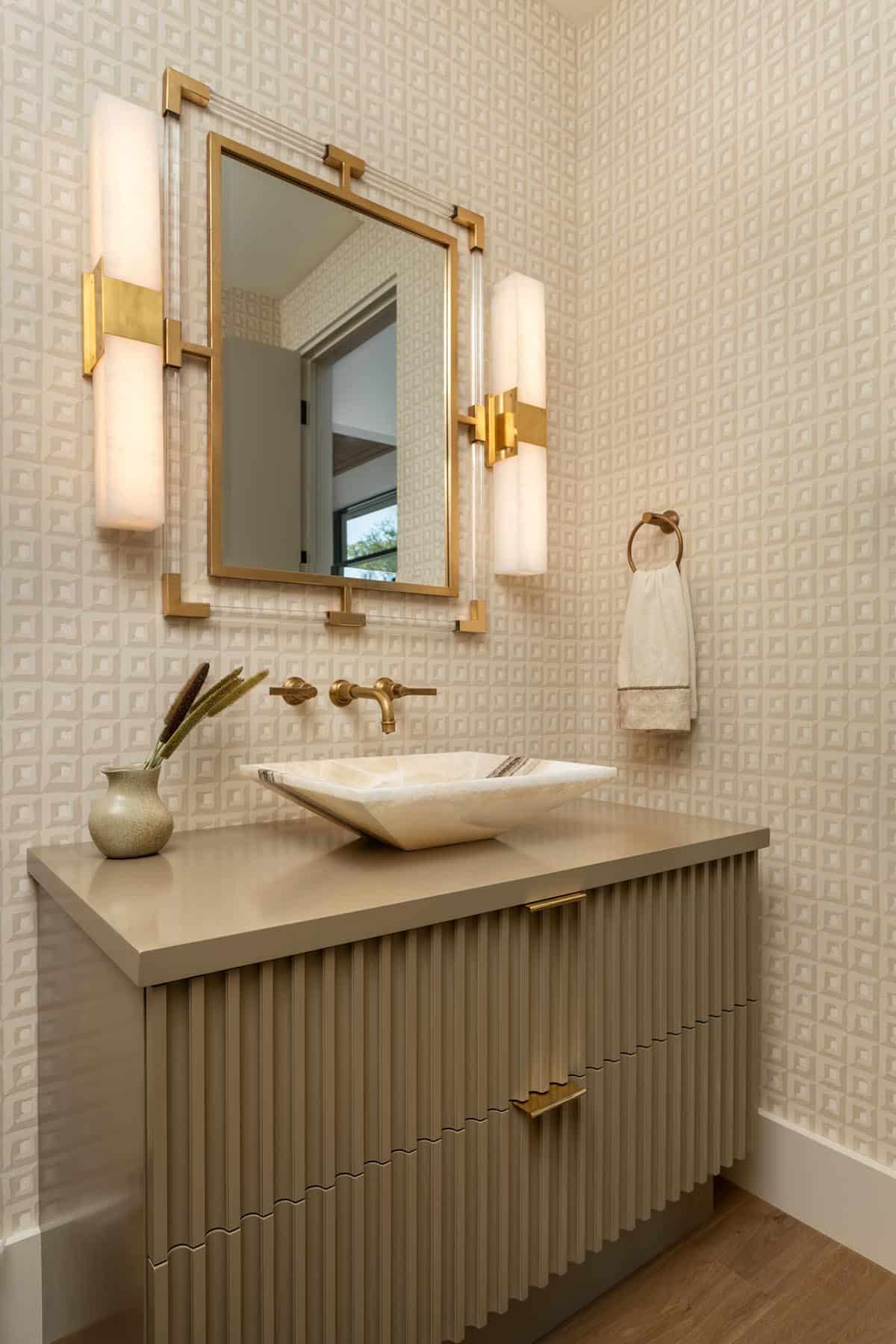 Powder room with fluted vanity, stone vessel sink, gold mirror, and patterned wallpaper