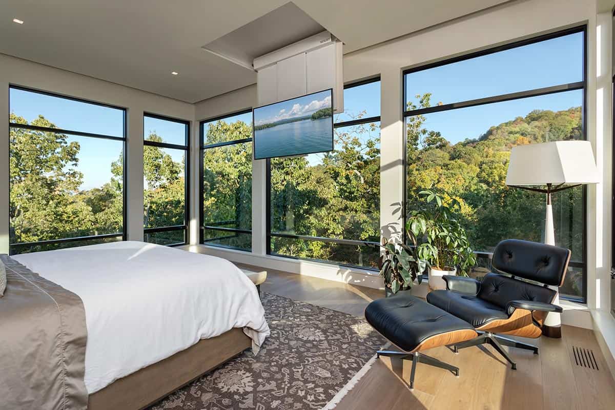 Primary bedroom with ceiling-mounted TV, Eames lounge chair, and treetop views