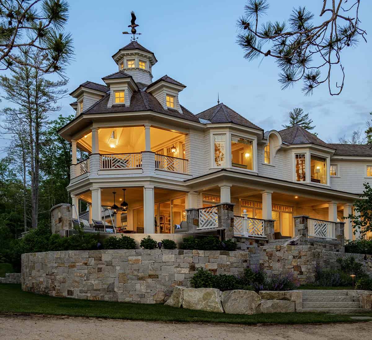 Twilight exterior of shingle-style lake house with turret and wraparound porch above stone retaining wall
