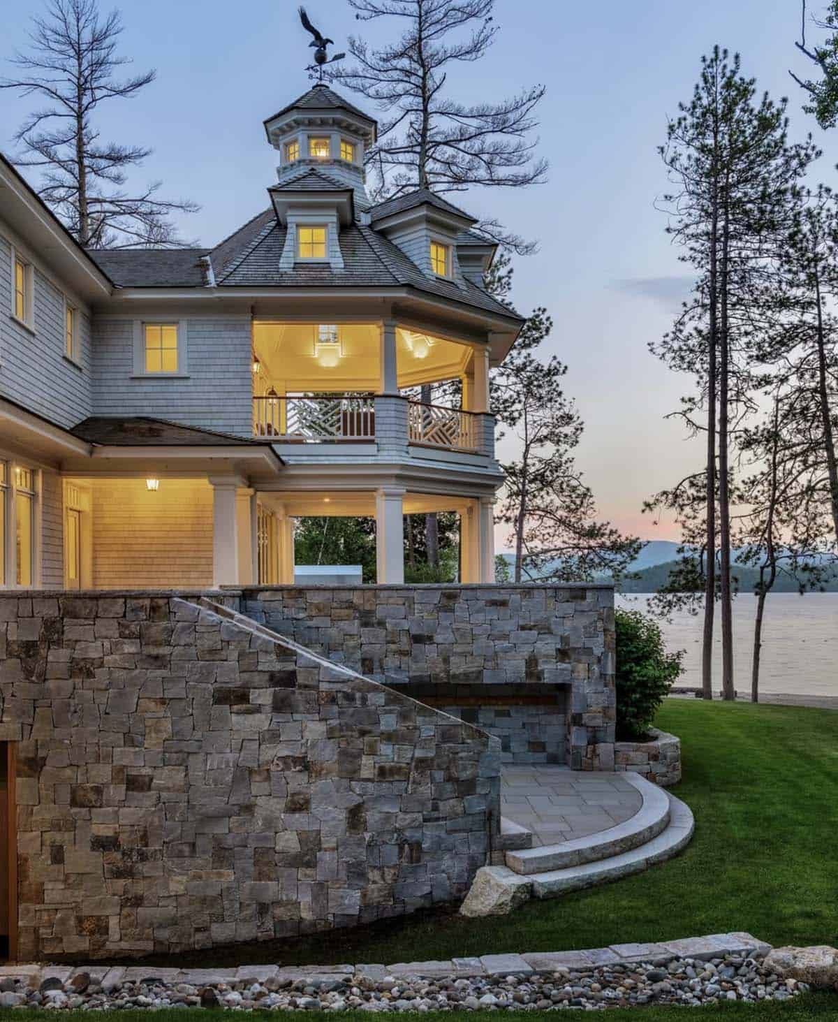 Shingle-style turret and covered porch overlooking lake at dusk, mountains in distance