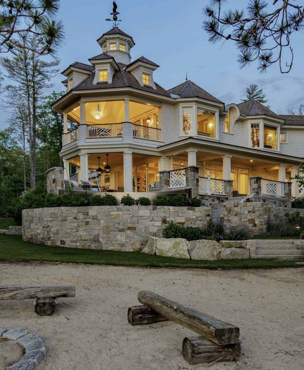 Lake house exterior at dusk with stacked porches, stone wall, and log benches around fire pit