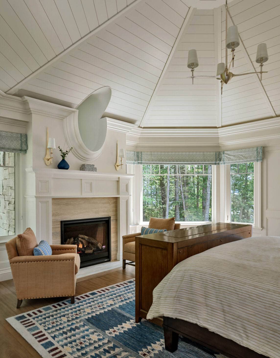 Bedroom with vaulted shiplap ceiling, fireplace, oval mirror, and bay window overlooking trees