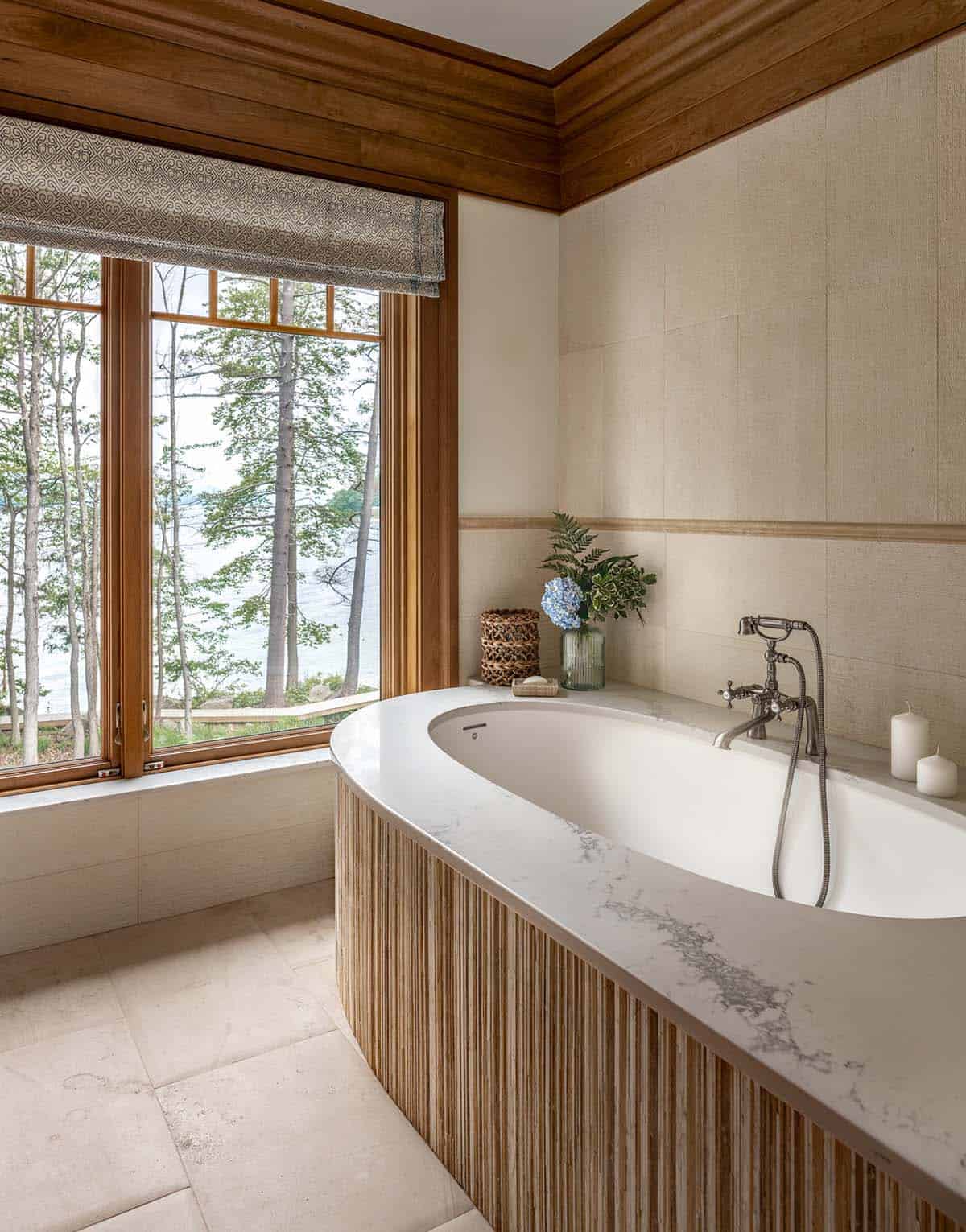 Freestanding tub with fluted wood surround beneath large window framing lake and trees