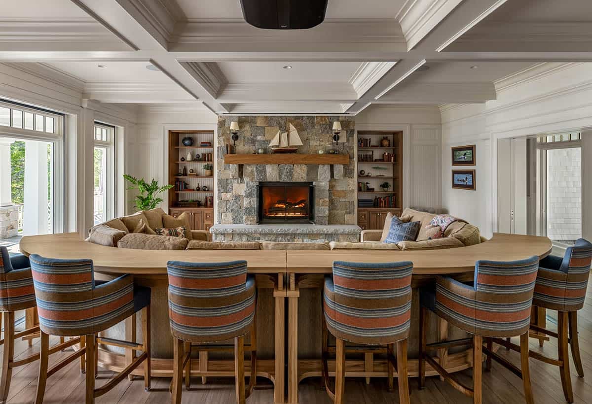 Curved oak bar with striped barstools facing a stone fireplace and built-in bookshelves