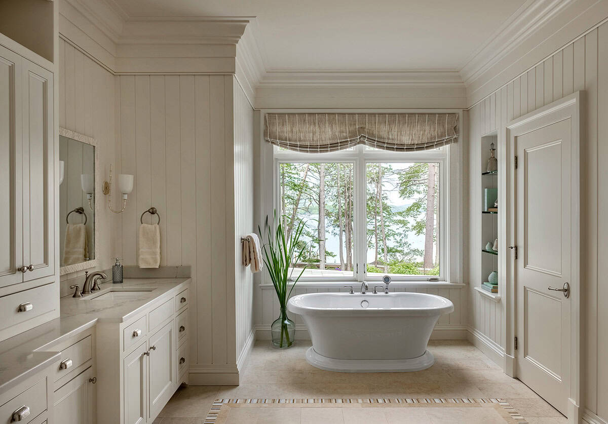 Freestanding white tub beneath picture window framing trees and water