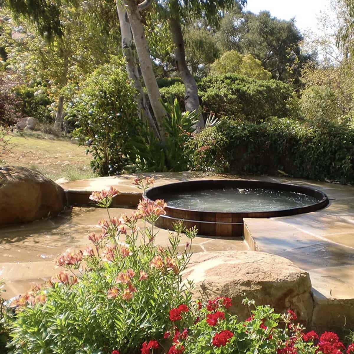 A round wood barrel hot tub set into a natural stone patio surrounded by boulders, flowering plants, and mature trees