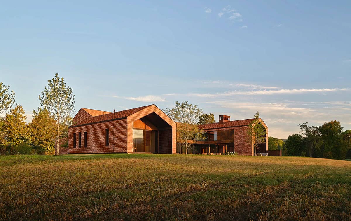 Full compound exterior at golden hour showing cedar-clad barn forms and Corten steel connections across meadow