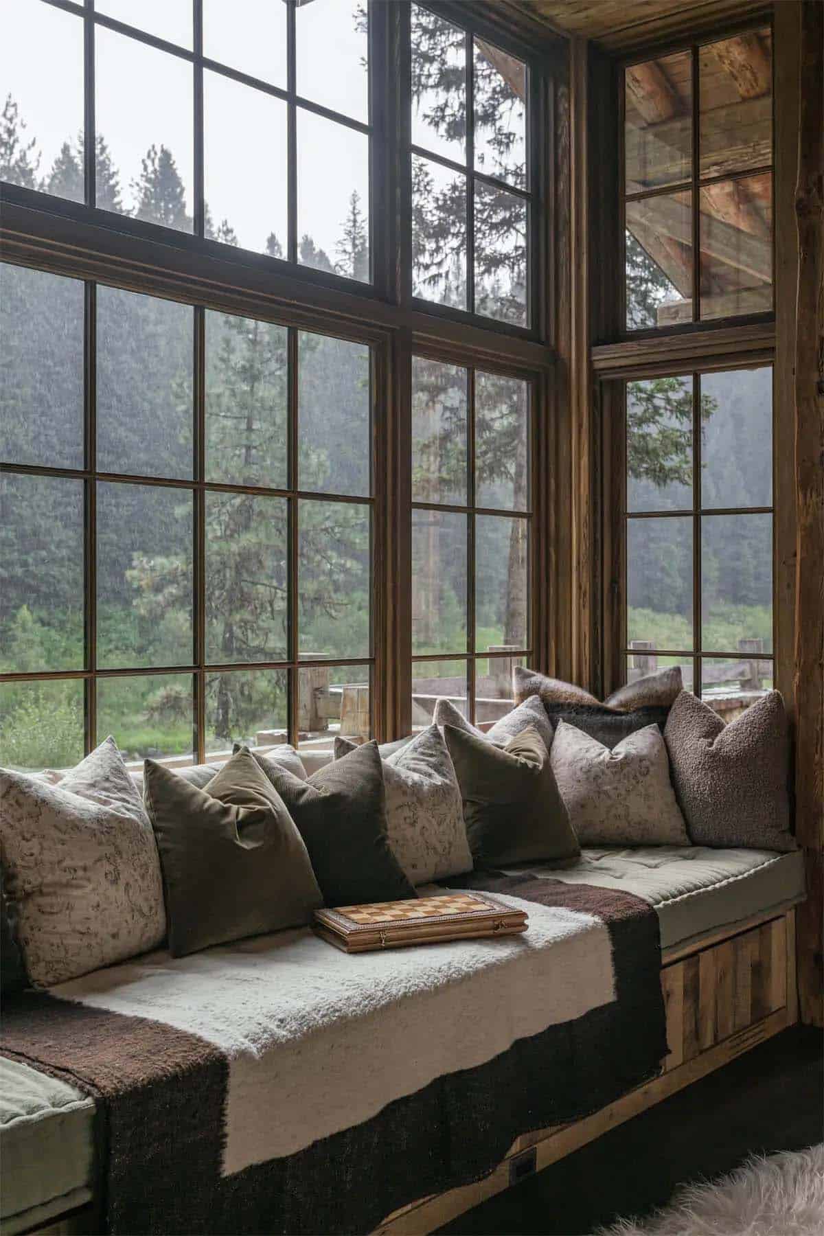 Window seat with layered pillows overlooking rainy forest, wooden beamed corner