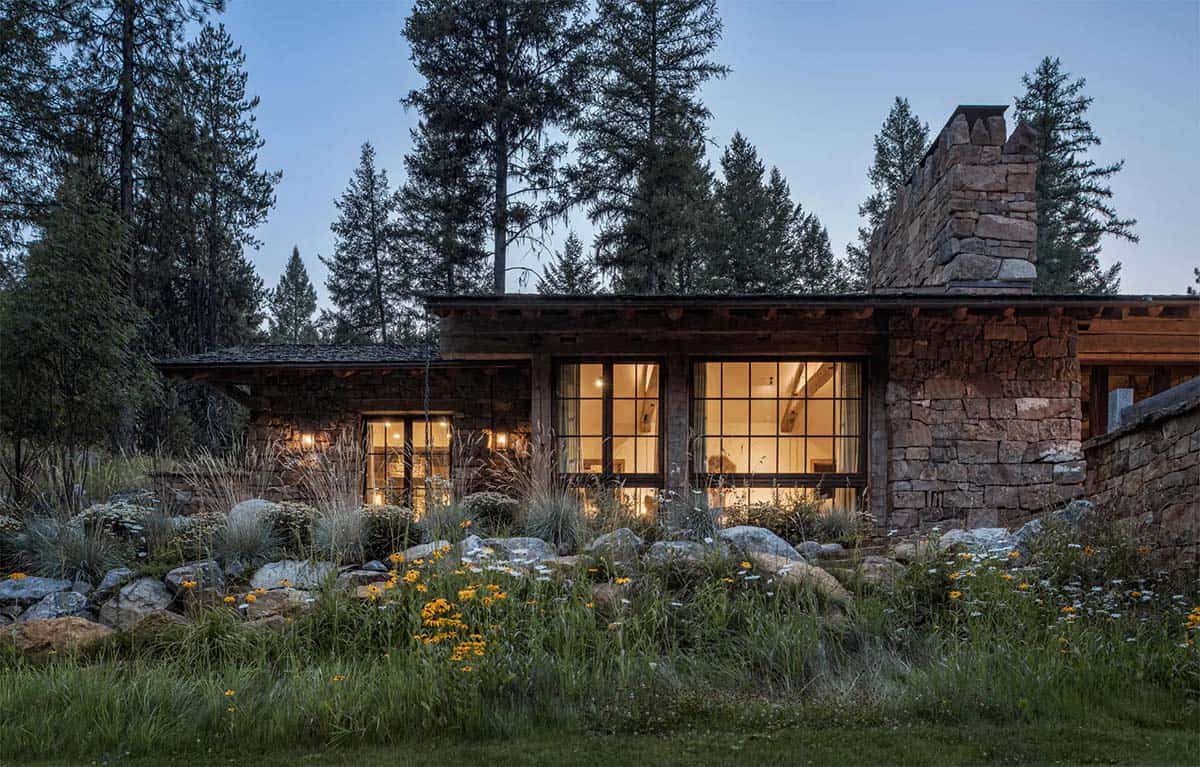 Stone and timber cabin exterior at dusk surrounded by wildflowers and pines