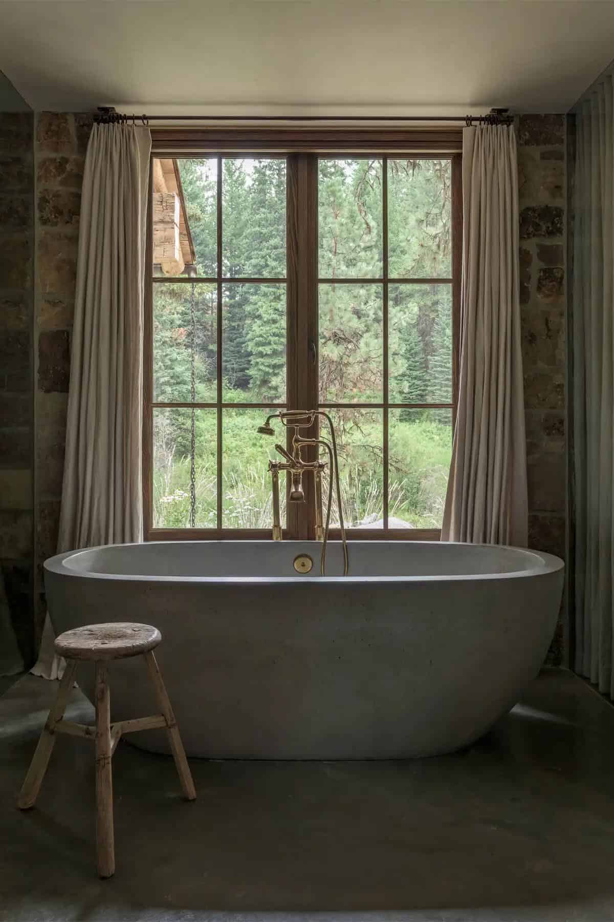 Concrete bathtub with brass faucet facing forest-view window