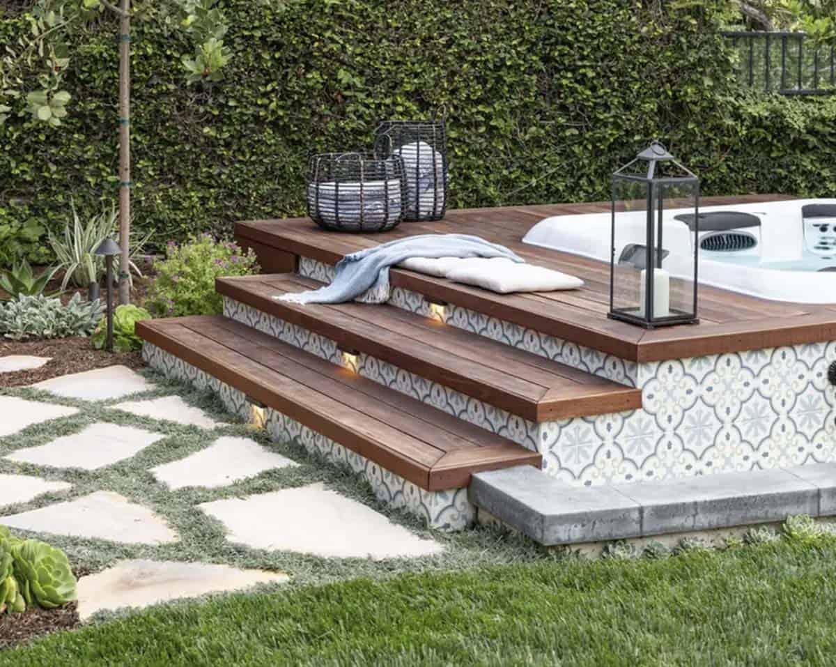 A raised wood deck hot tub with Spanish tile trim, lit wood steps, and a flagstone path in an Orange County backyard