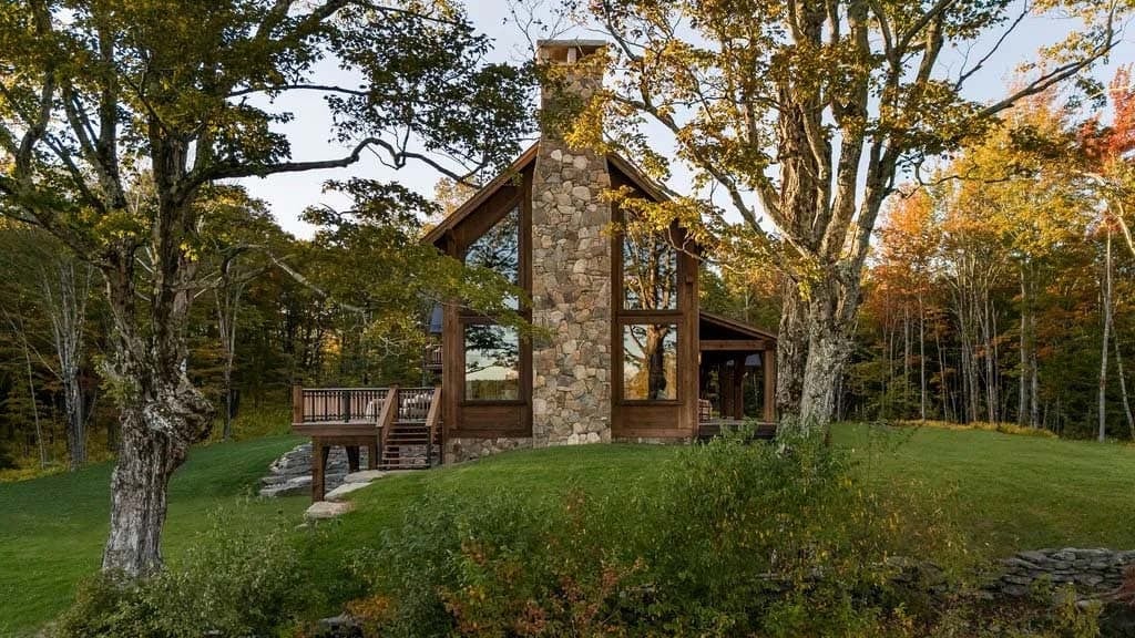 Timber frame mountain home exterior with stone chimney, surrounded by fall foliage at dusk