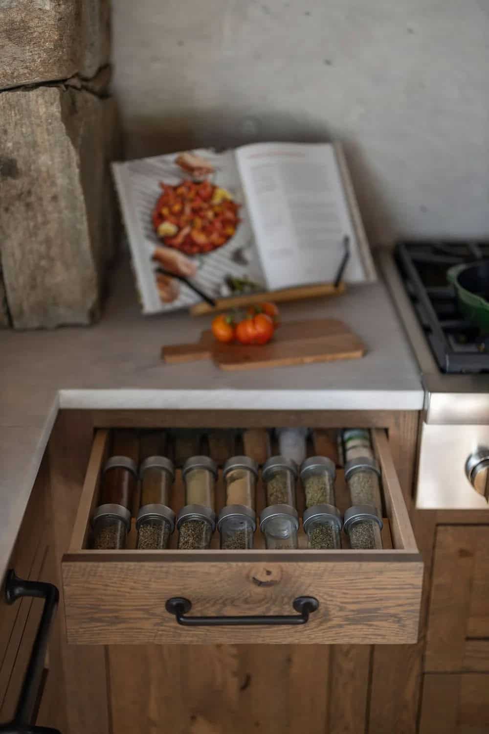 Open spice drawer with glass jars beside kitchen stove and cookbook