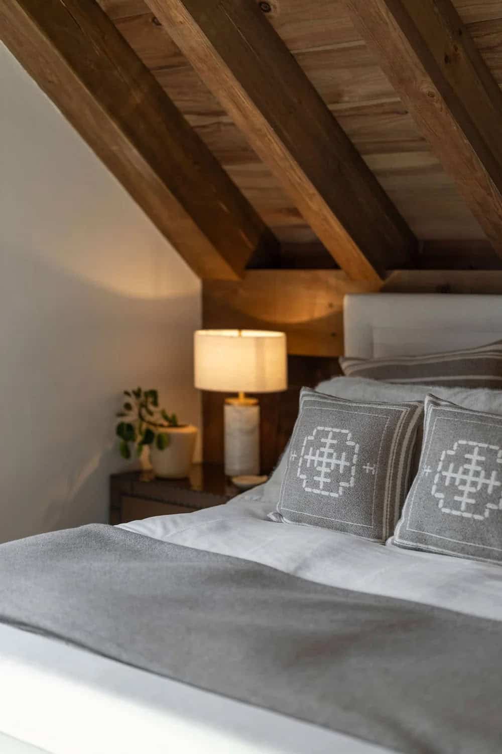 Cozy bedroom nook under sloped wood ceiling with bedside lamp and pillows