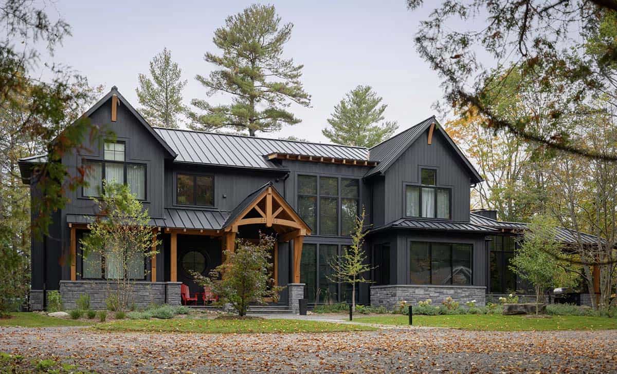 Dark exterior of a transitional cottage with black board-and-batten siding, timber frame entry, and metal roof surrounded by pine trees