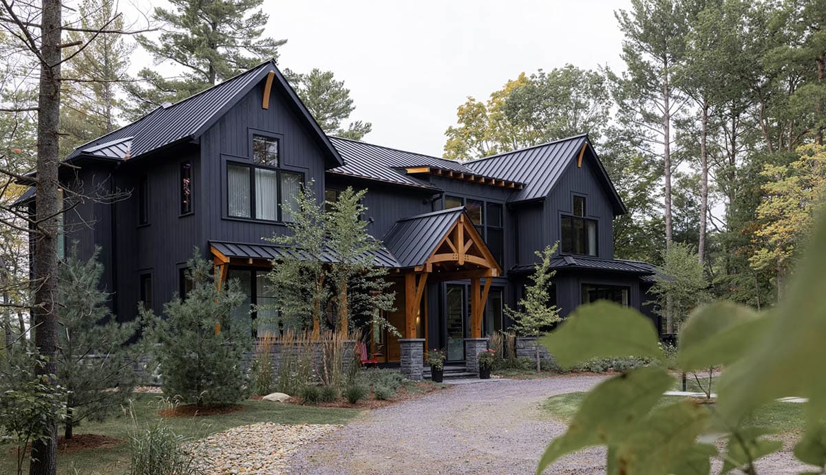 Angled view of dark cottage exterior with timber brackets, steep gabled rooflines, and mature tree landscaping