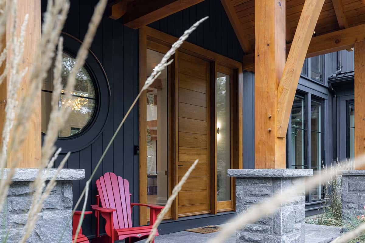 Close-up of cottage entryway with natural wood door, round porthole window, red Adirondack chair, and timber frame porch