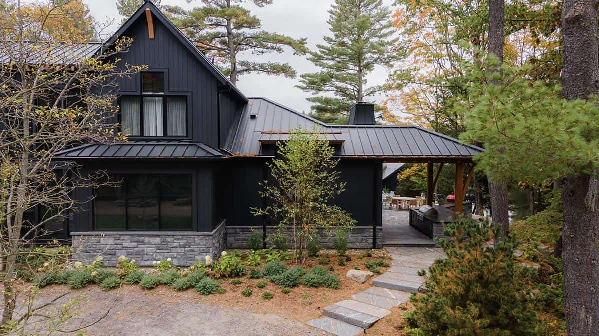 Side view of black cottage exterior with stone chimney, covered outdoor kitchen pavilion, and flagstone path