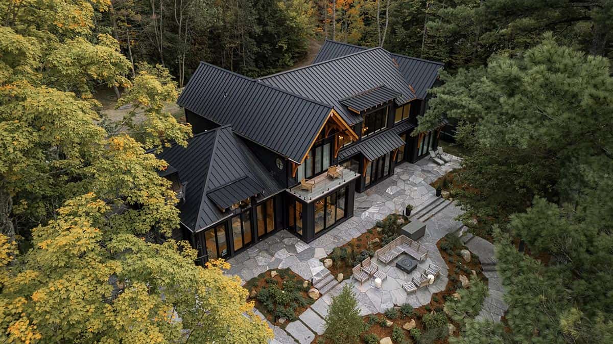 Aerial view of black cottage roofline with stone patio and fire pit seating area nestled in autumn forest