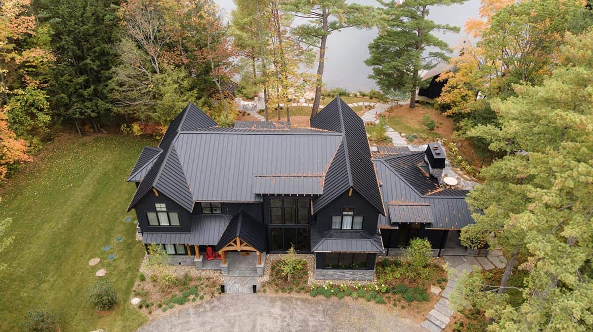 Overhead view of black cottage with complex multi-gabled roofline, lake visible through autumn trees