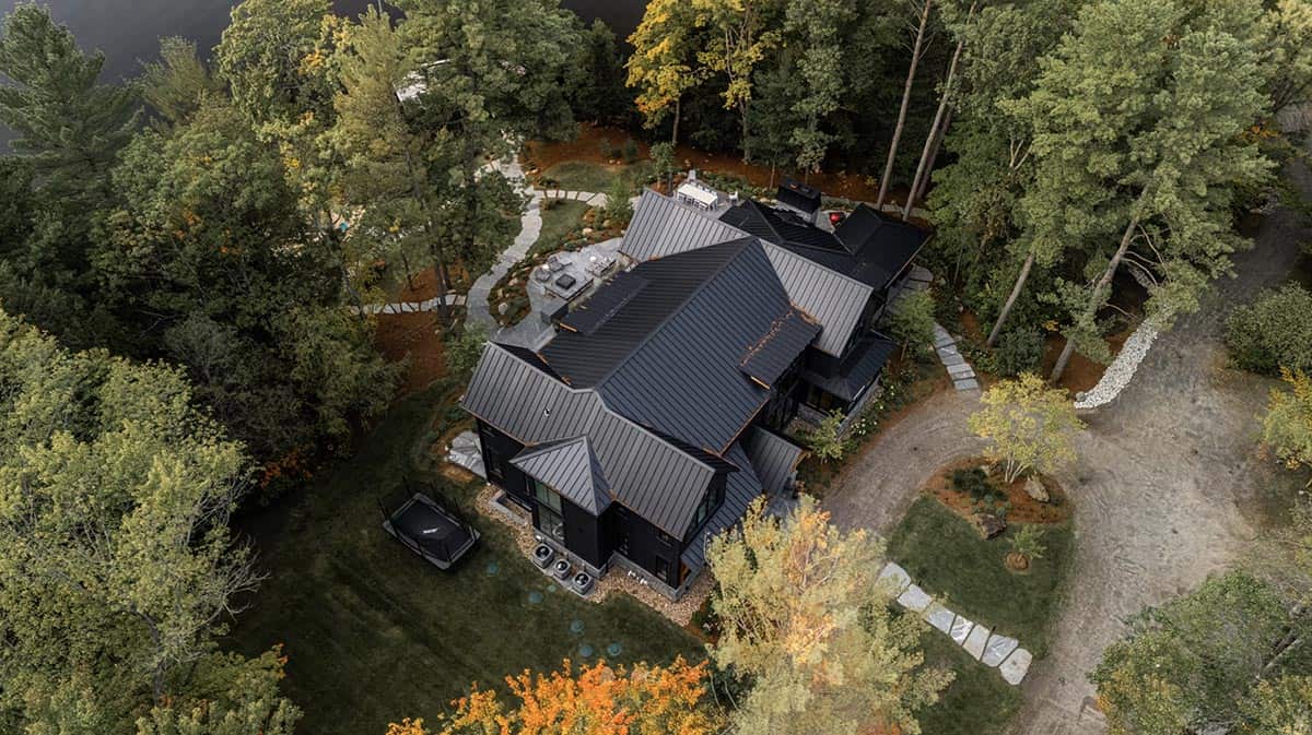 High aerial view of black cottage complex surrounded by dense forest with lake glimpsed through the canopy
