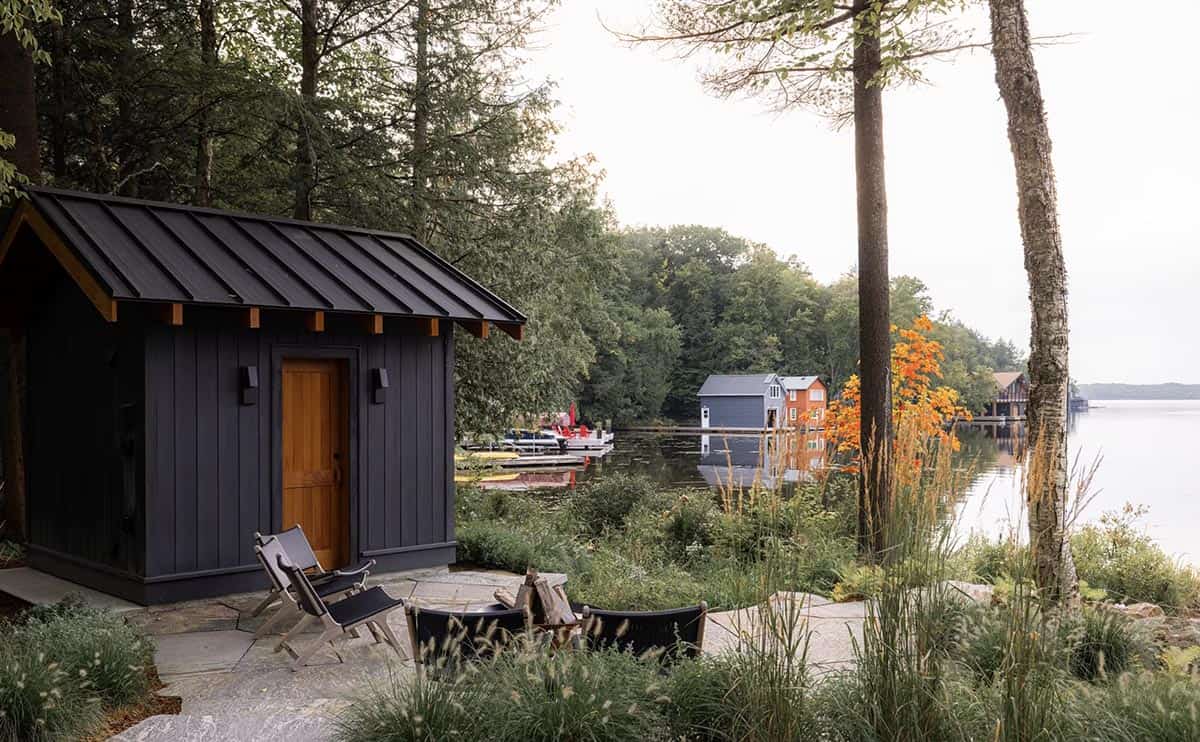 Small black lakeside sauna cabin with wood door, fire pit, and Muskoka lake in the background