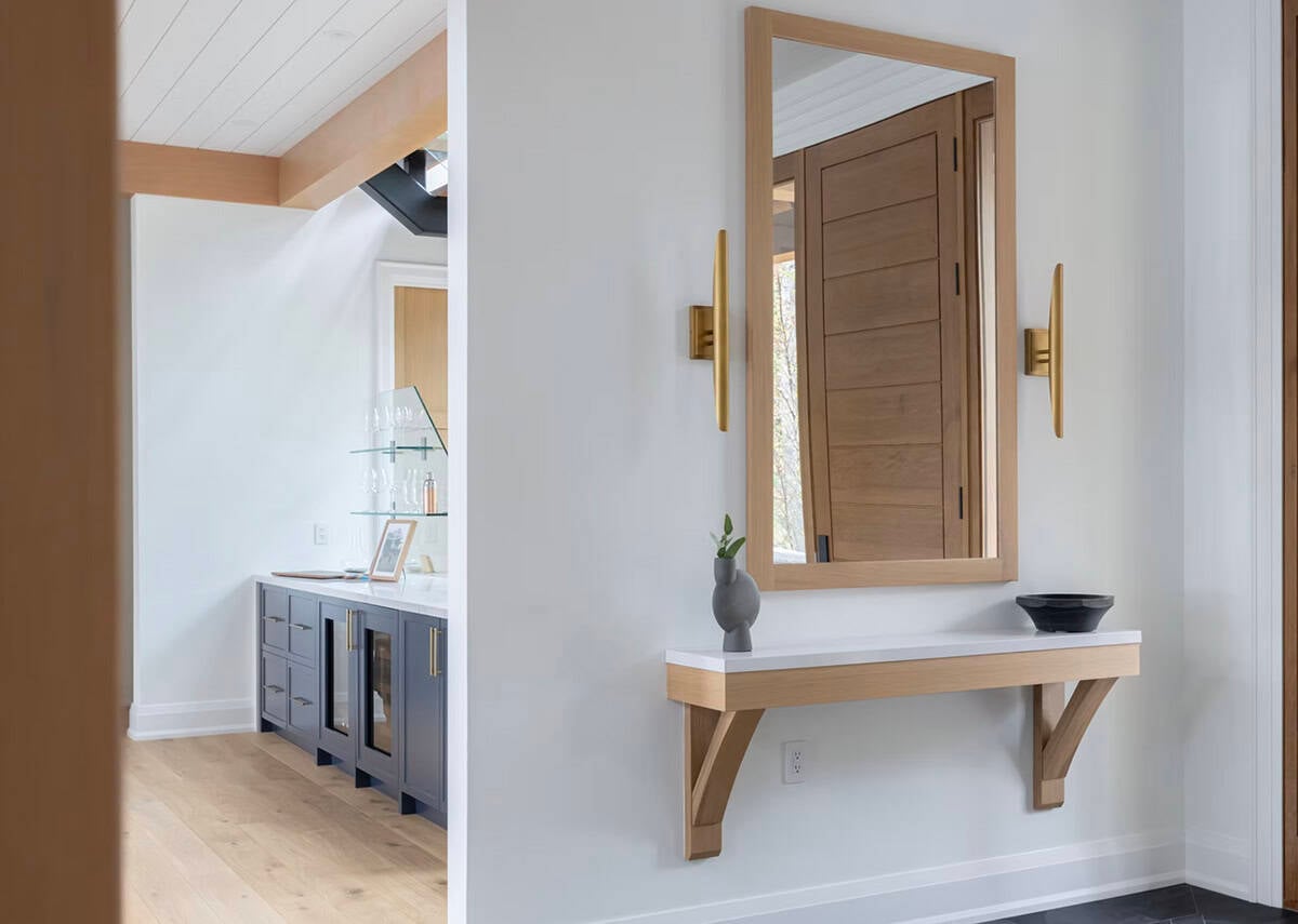 Bright cottage entryway with oak-framed mirror, floating shelf, brass sconces, and navy butler's pantry beyond