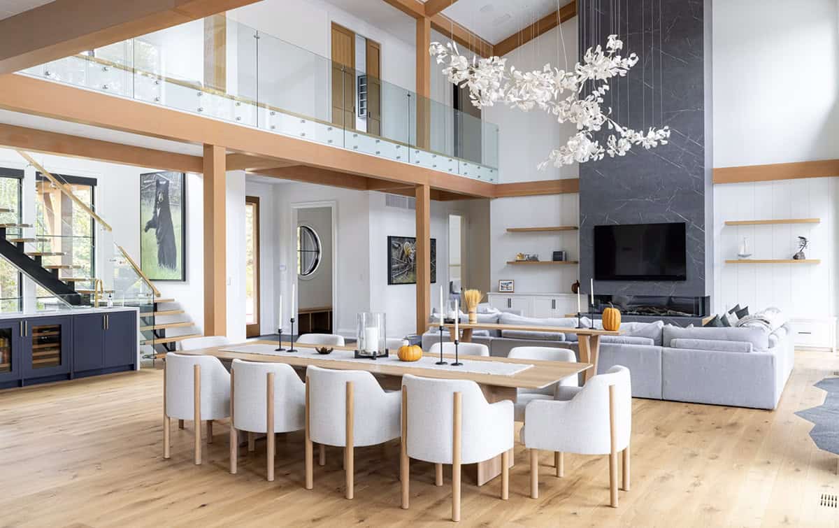 Open-plan great room with double-height ceiling, white floral chandeliers, timber beams, dining table, and stone fireplace beyond
