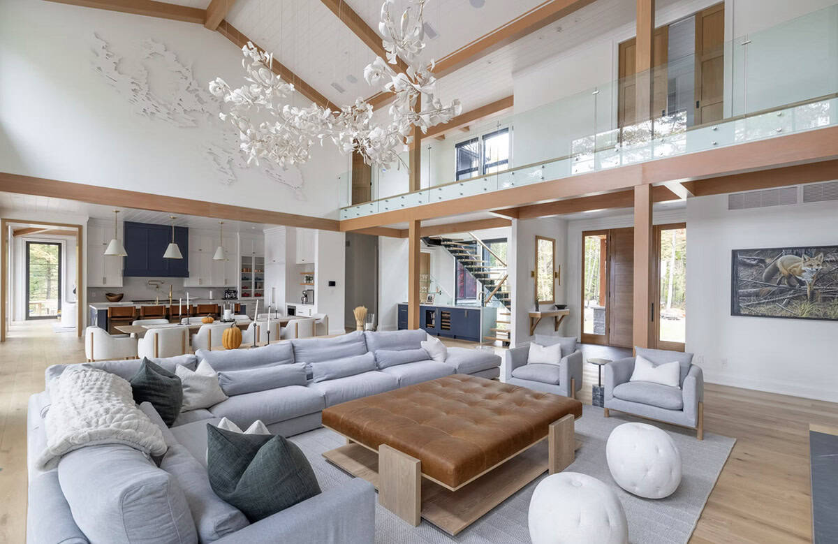 Wide-angle view of open-plan cottage interior with grey sofas, leather ottoman, lake map wall art, and kitchen beyond