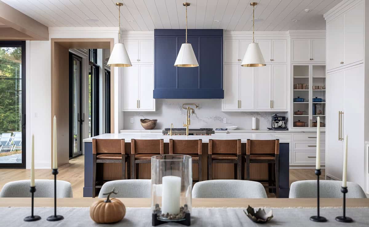 Kitchen island with leather stools viewed from dining table, navy range hood and white cabinetry with brass hardware