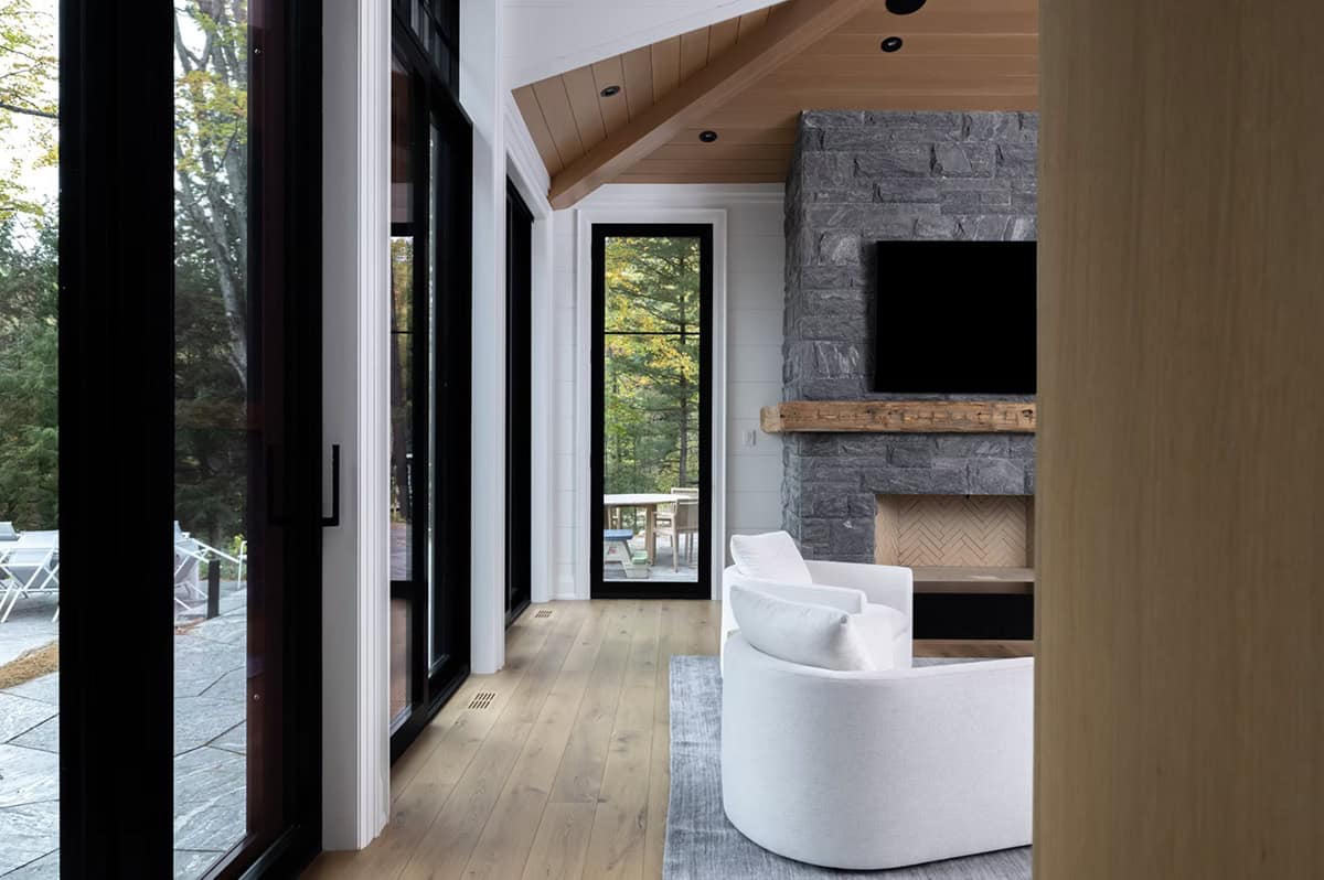 Bright sunroom with floor-to-ceiling black windows, vaulted cedar ceiling, and stone fireplace with reclaimed wood mantel