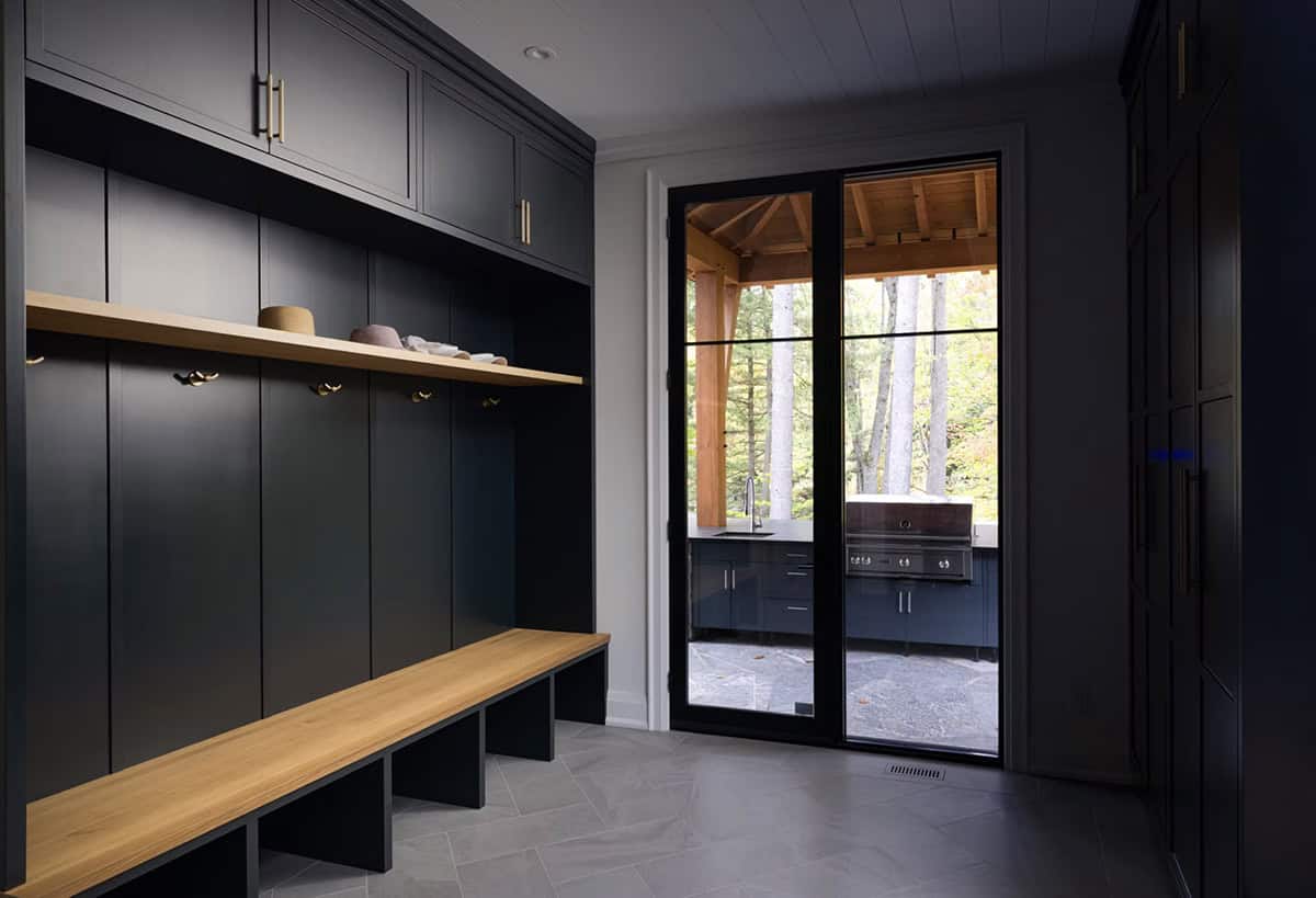 Dark charcoal mudroom with built-in lockers, oak bench, brass hooks, and glass door to covered outdoor kitchen pavilion