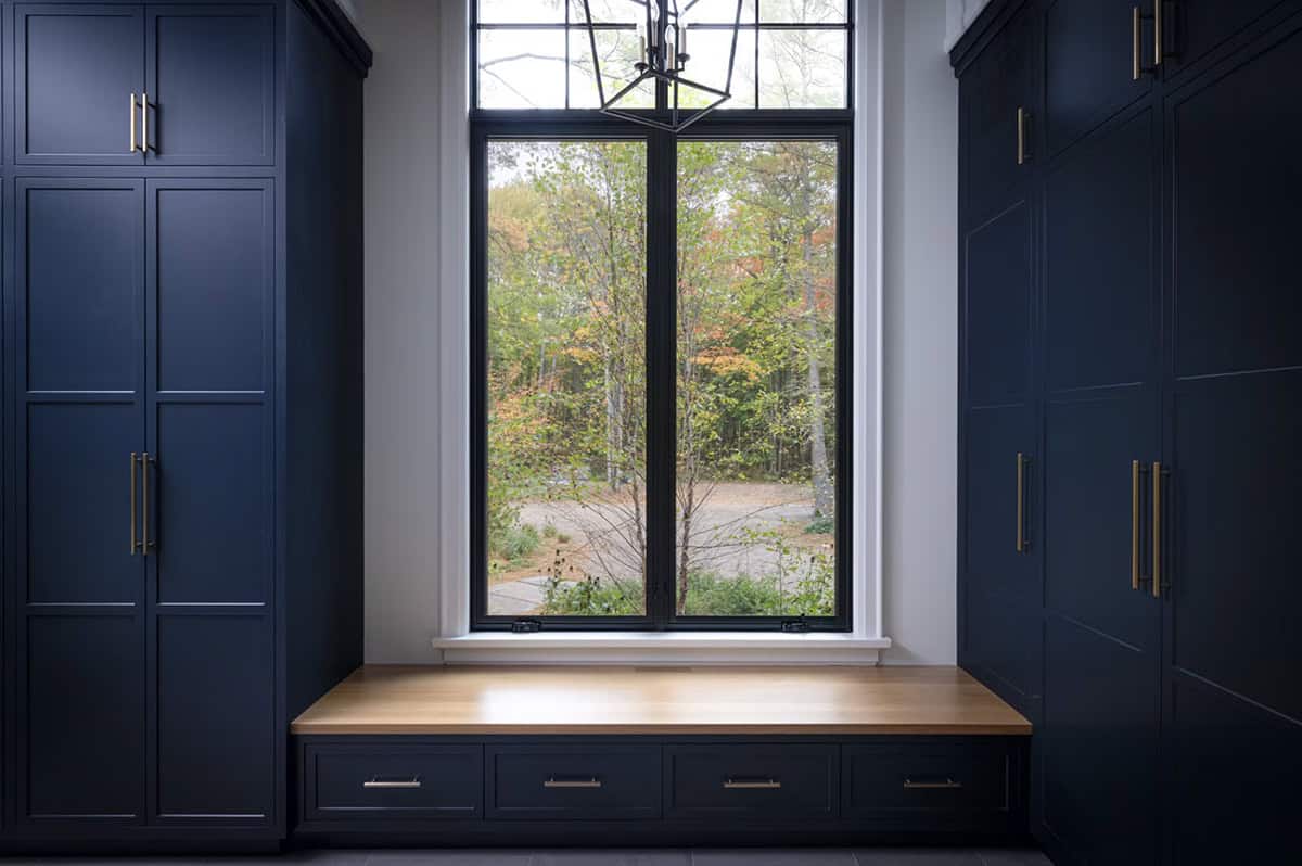 Navy mudroom with built-in storage cabinets, oak window bench with drawers, and black-framed window overlooking forest
