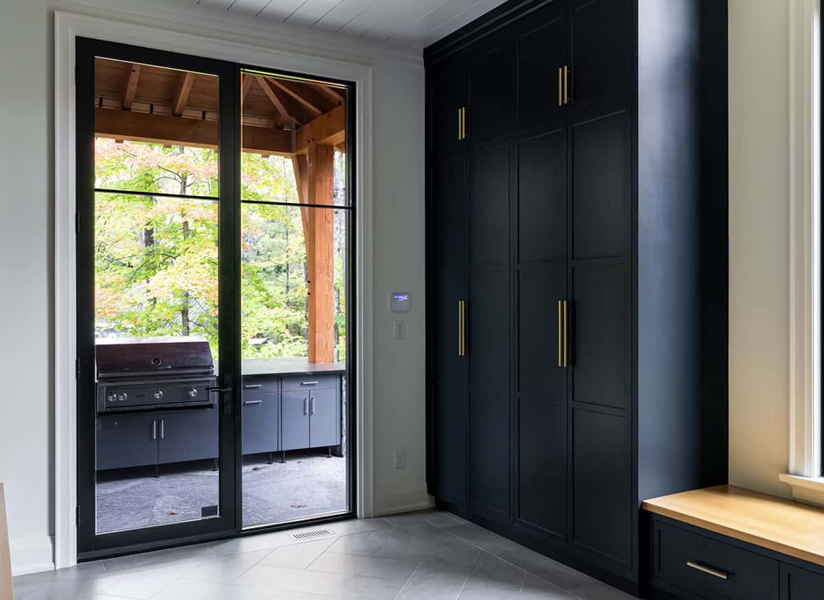 Dark mudroom with floor-to-ceiling navy cabinetry, brass handles, and glass door view of outdoor kitchen under timber pergola