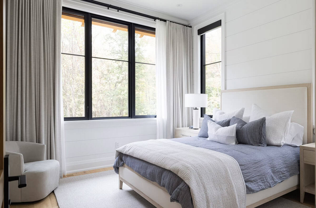 Serene guest bedroom with blue linen bedding, upholstered headboard, shiplap accent wall, and corner black-framed windows with forest views