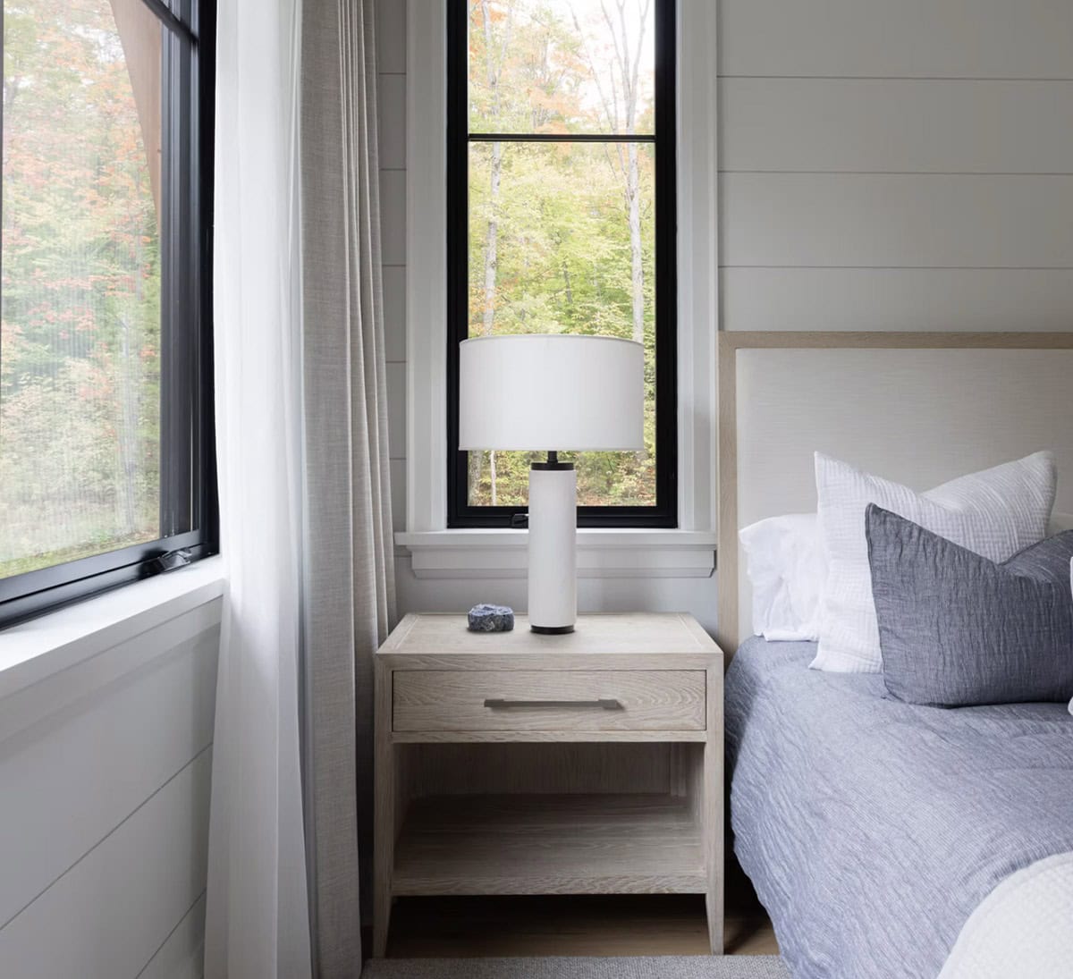 Close-up of oak nightstand with white lamp beside blue-sheeted bed and black-framed window overlooking fall foliage