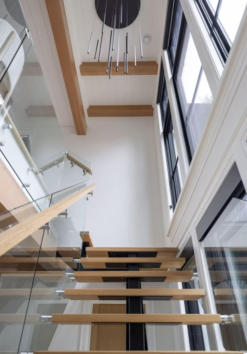 Looking down open-tread oak staircase with glass railings, black steel spine, and modern rod chandelier above