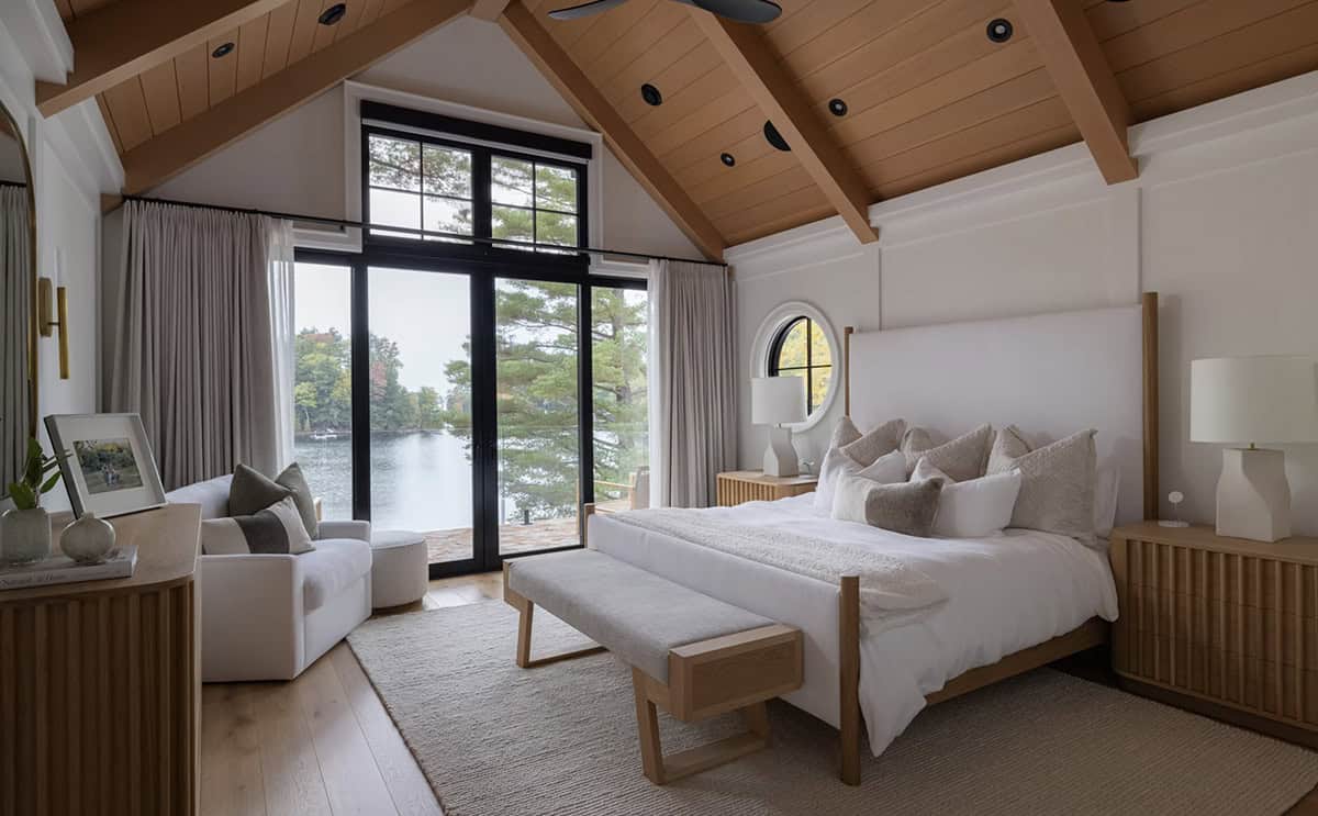Primary bedroom with vaulted cedar ceiling, timber beams, white king bed, porthole window, and floor-to-ceiling lake view