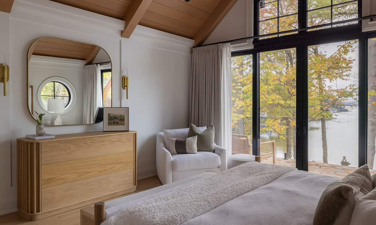 Primary bedroom corner with fluted oak dresser, arched gold mirror, white armchair, and open glass door to lakeside balcony
