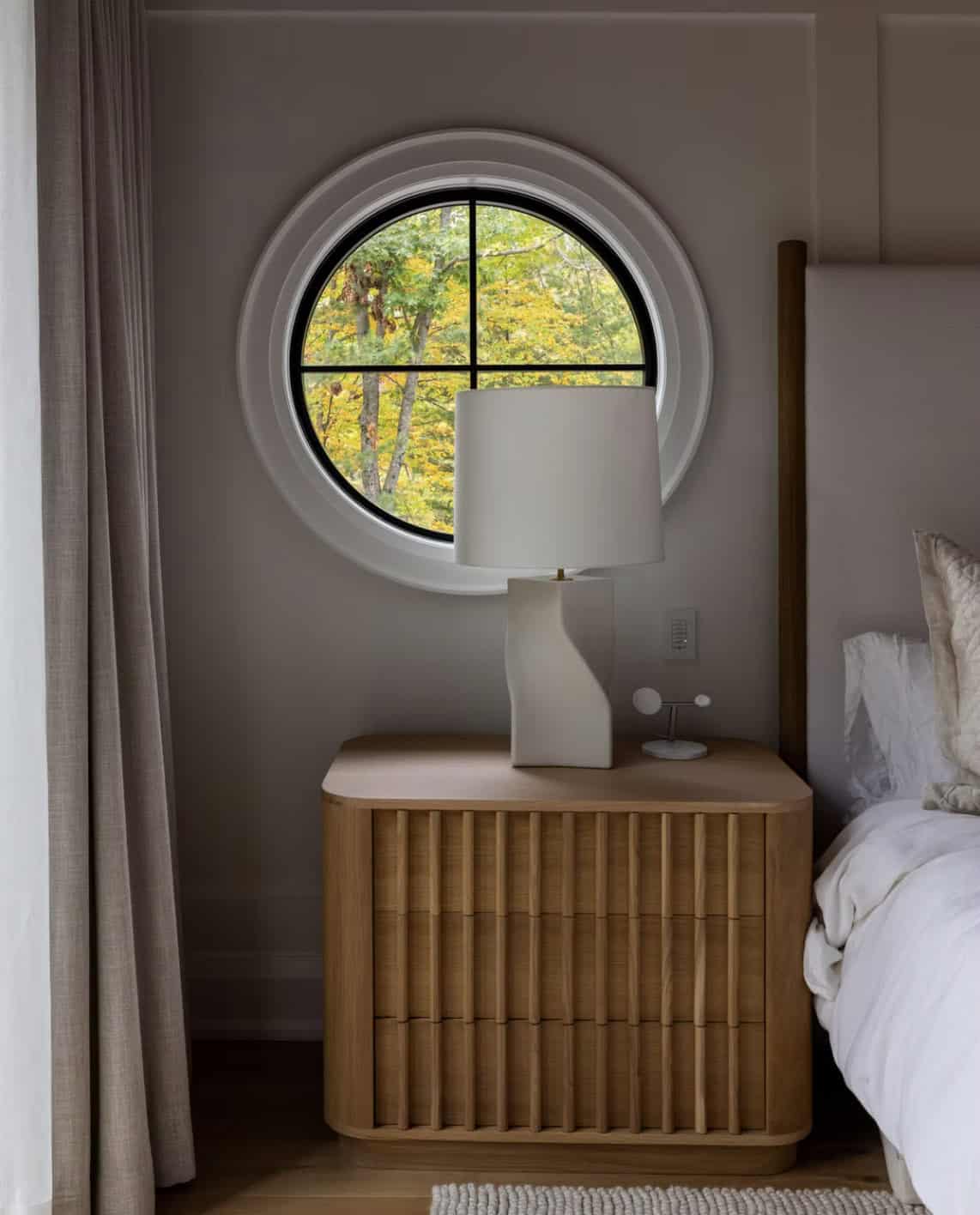 Fluted oak nightstand with sculptural white lamp centered under round porthole window framing fall foliage