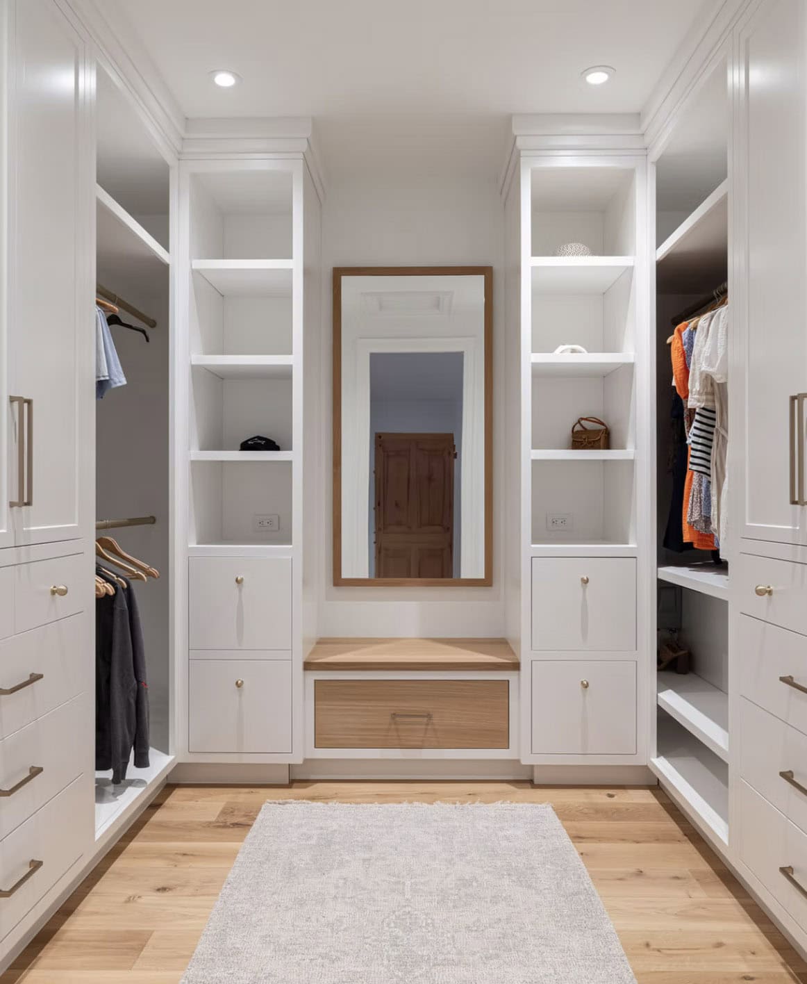 Walk-in closet with white built-in cabinetry, brass hardware, oak vanity bench, and full-length mirror