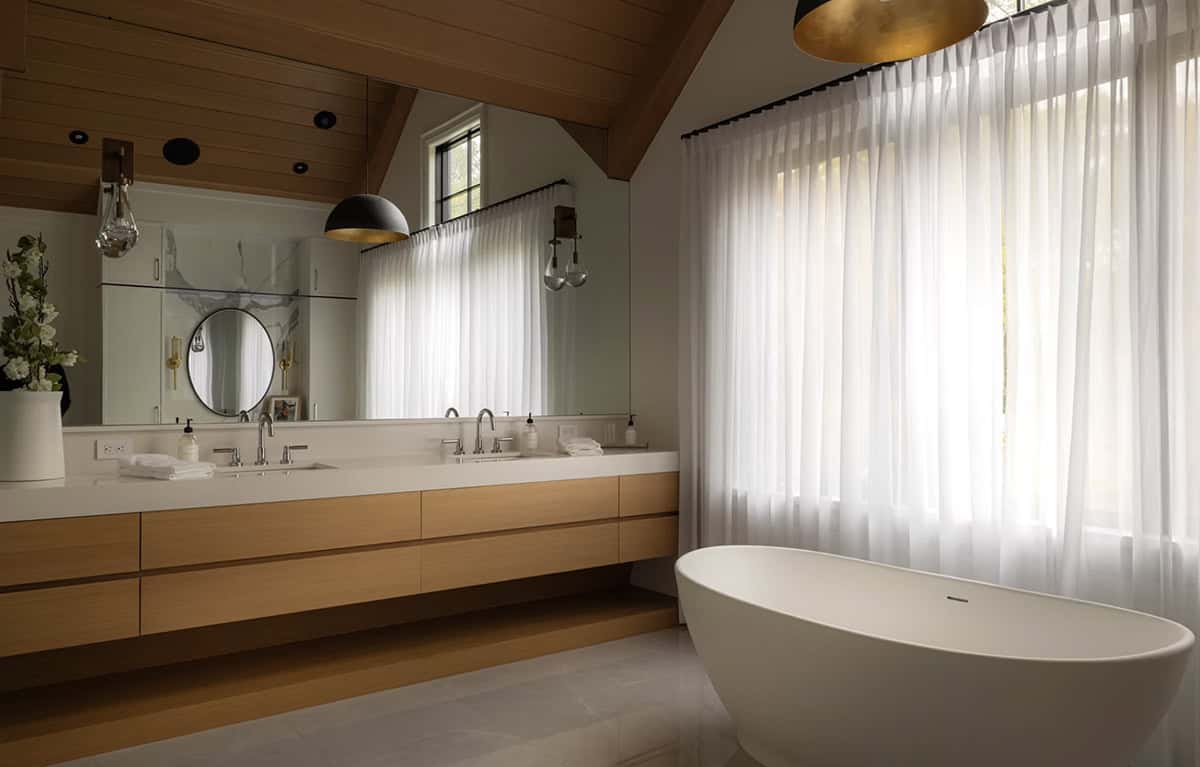 Primary ensuite with freestanding soaker tub beside sheer-draped window, oak double vanity, and vaulted cedar ceiling