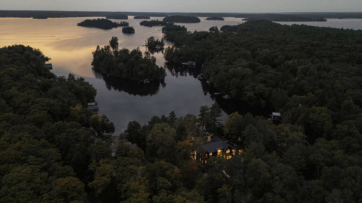 Sweeping aerial view of glowing Muskoka cottage at dusk nestled in forest beside a calm island-dotted lake