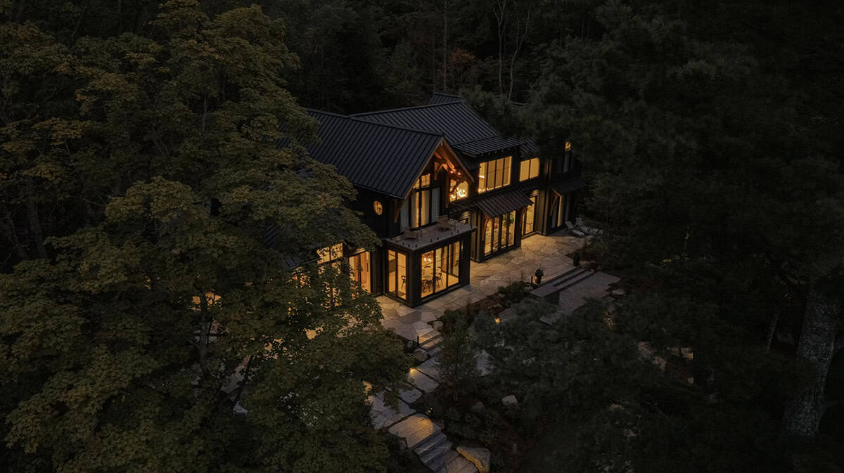 Dusk aerial view of black cottage glowing from within, stone patio illuminated, set in dense forest canopy