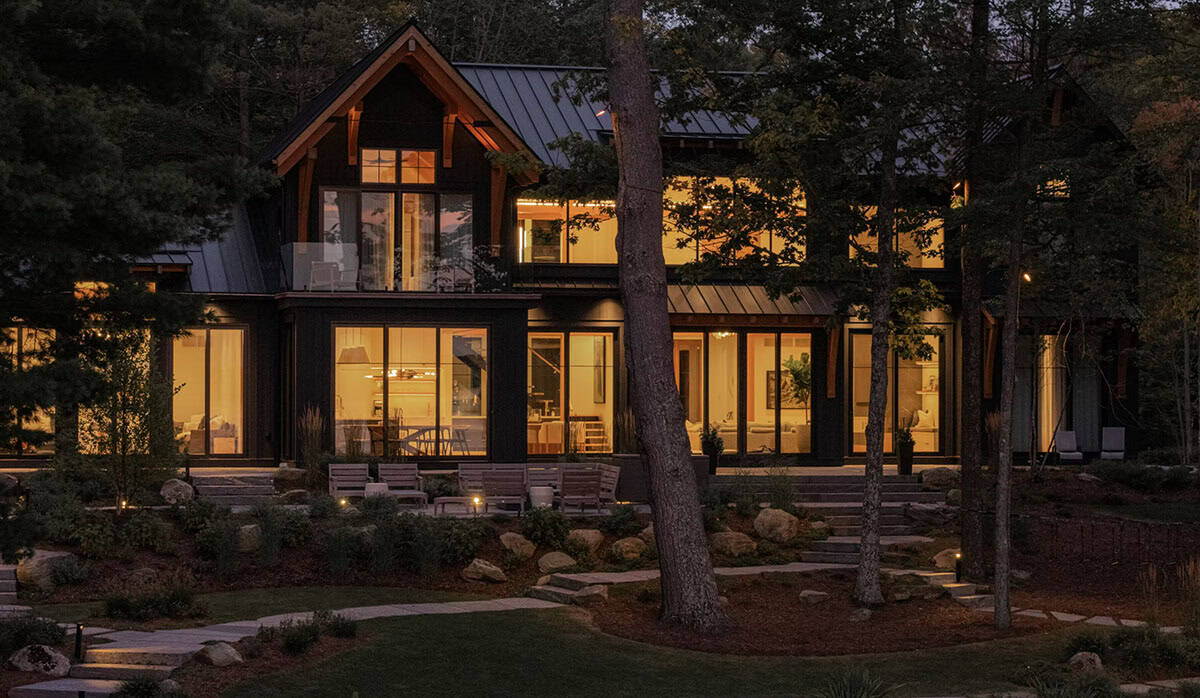 Twilight view of lit cottage lakeside facade with timber-framed gable, balcony, wraparound glazing, and stone patio