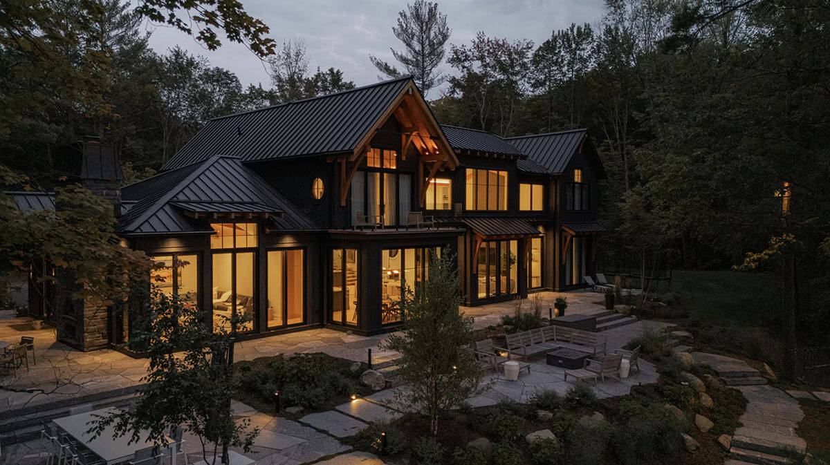Dusk exterior of black Muskoka cottage with glowing windows, timber-framed gable, stone patio, and fire pit seating