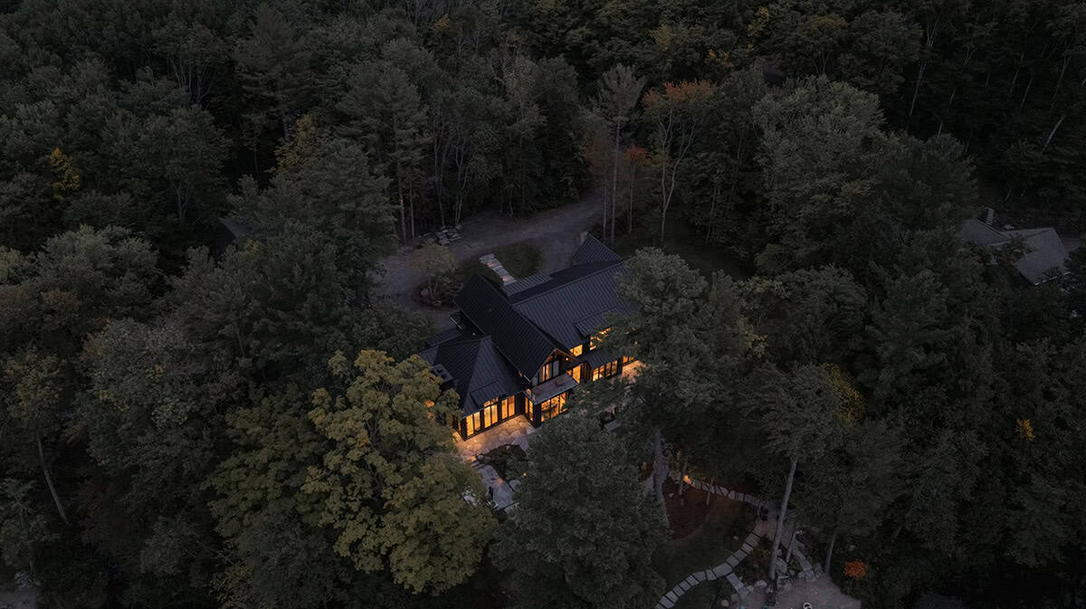 Nighttime aerial of illuminated black cottage tucked deep in Muskoka forest with winding stone path visible below