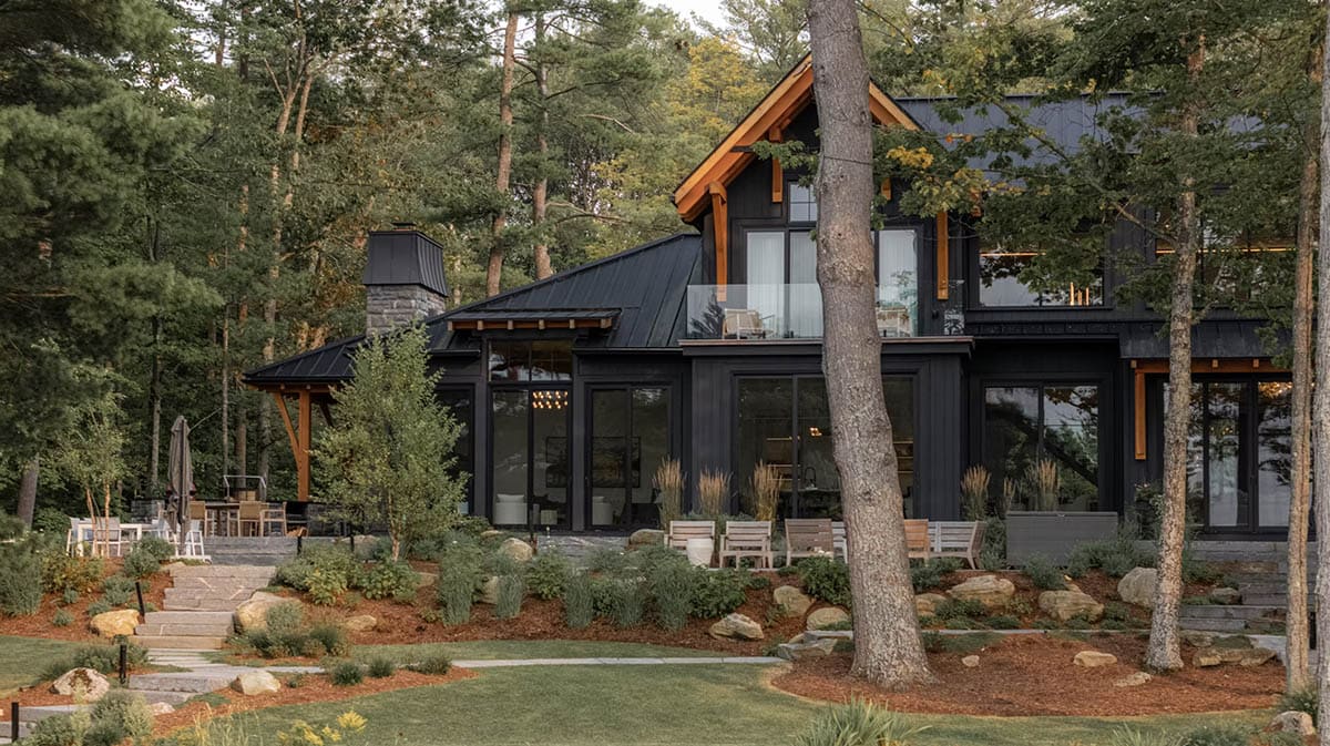 Lakeside rear facade of black cottage with timber brackets, balcony, wraparound glazing, stone patio, and natural landscaping