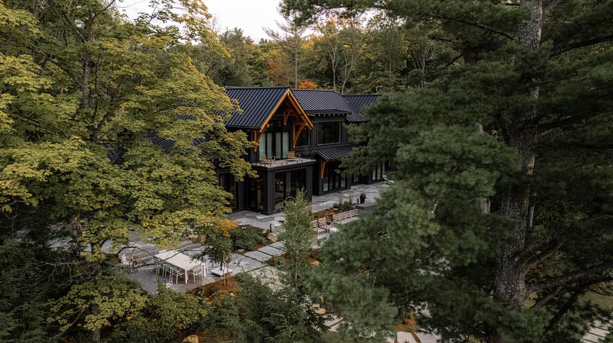 Aerial view of black cottage lakeside facade with timber-framed gable, stone terrace, and early fall foliage canopy