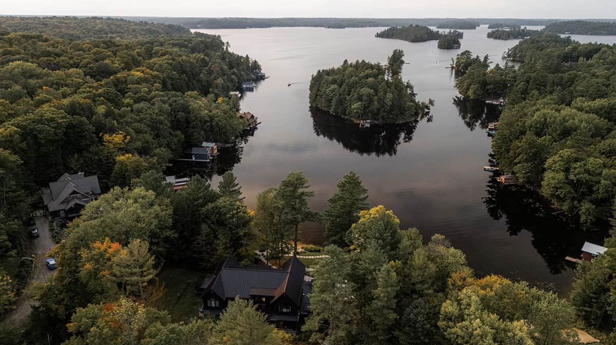 Wide aerial view of black Muskoka cottage on calm tree-lined bay with forested islands and open lake beyond